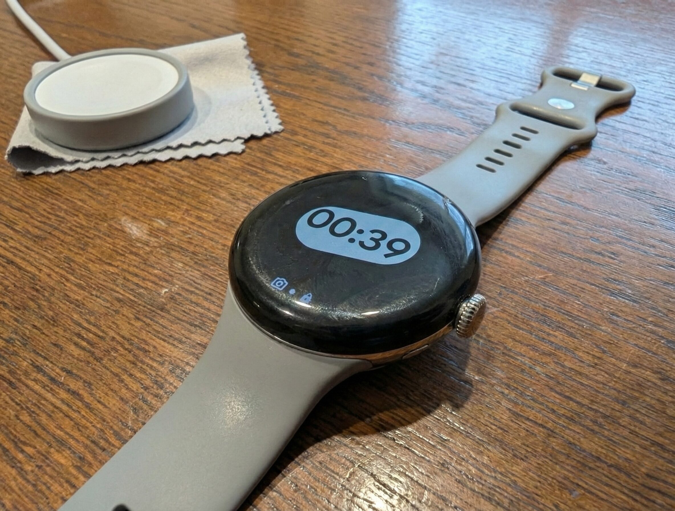 1.5年使用されたGoogle Pixel Watch 3。画面の右上に小さな傷があり、グレーのアクティブバンドには使用感によるテカリが見られる。本体の横には専用充電器とクリーニングクロスが木製の机の上に置かれている。